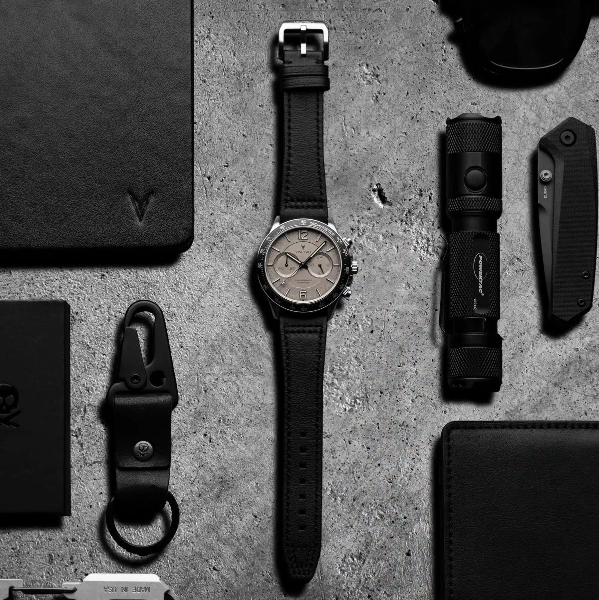 Black watch with leather strap on a textured gray surface, surrounded by other black items.