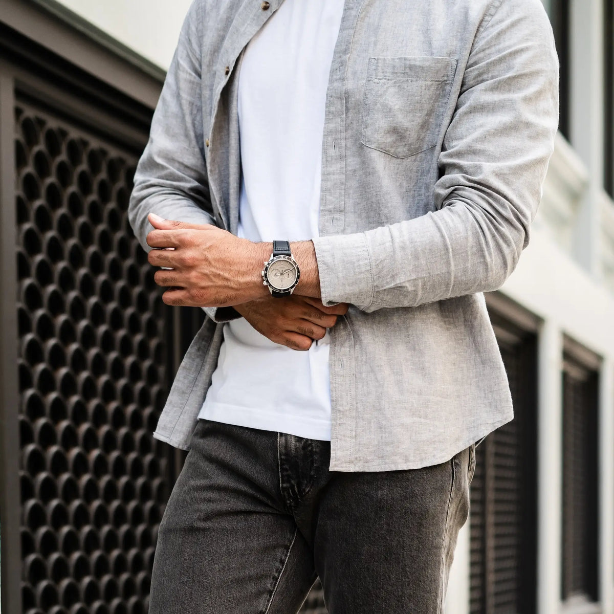 Person wearing a light gray jacket over a white shirt and dark jeans, standing against an urban background.