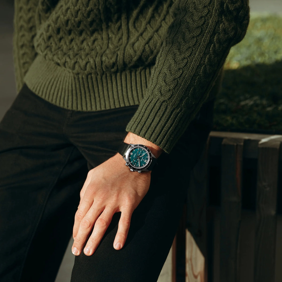 Person wearing a green knitted sweater and dark watch, sitting on a wooden bench.