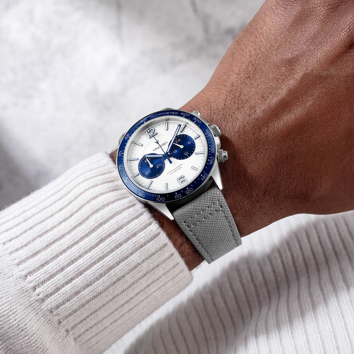 A wristwatch with a silver-toned case, white dial, blue bezel and subdials, and a gray textured strap, shown worn on a person's wrist over a white ribbed sweater.