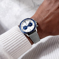 A wristwatch with a silver-toned case, white dial, blue bezel and subdials, and a gray textured strap, shown worn on a person's wrist over a white ribbed sweater.