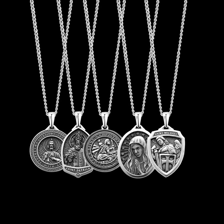 Five silver religious pendant necklaces with detailed saint engravings, including Saint Patrick, Saint Patrizio, Our Lady of Mount Carmel, Saint Michael, and Saint Monica, displayed against a solid black background.