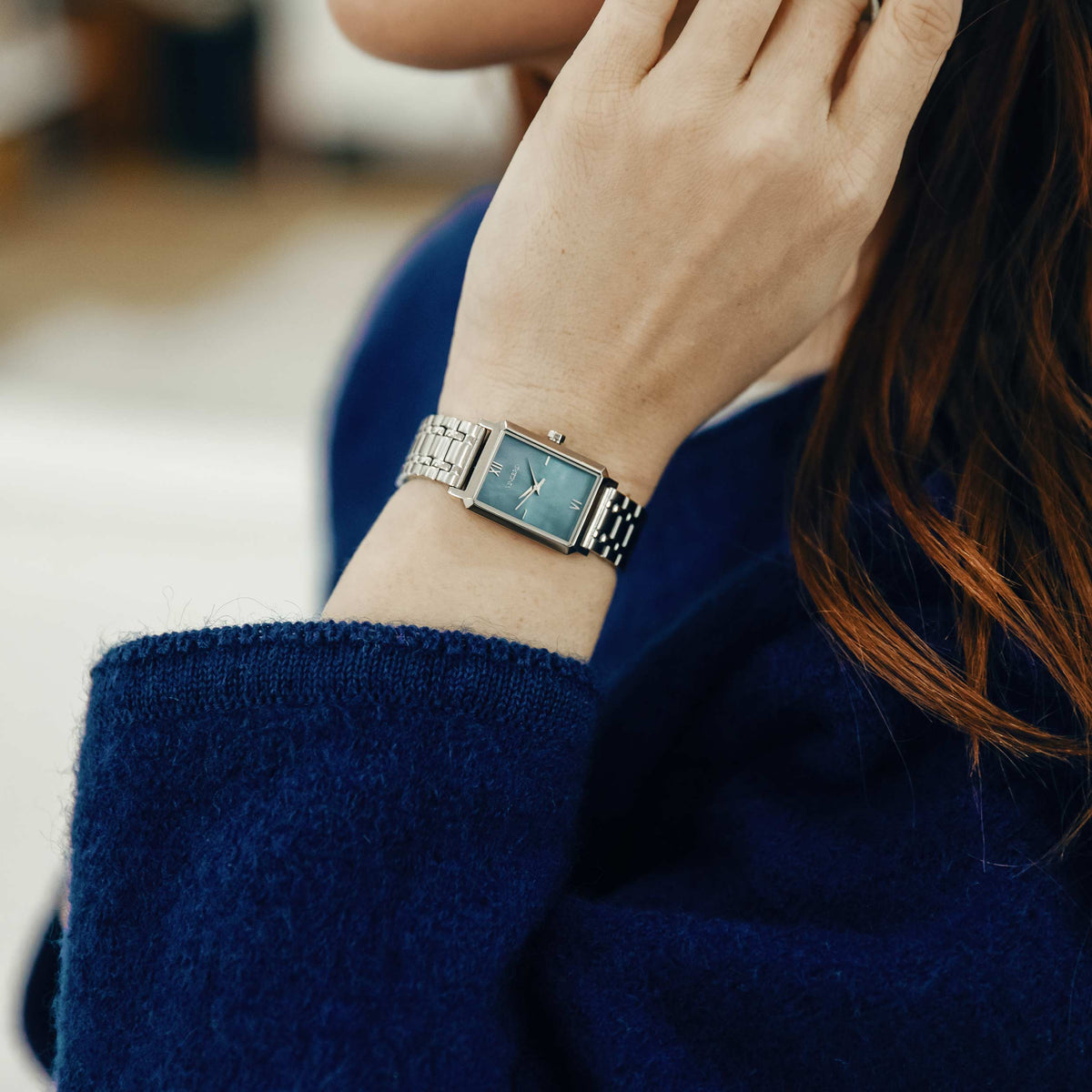 Close-up of a person wearing a blue dress with a matching watch on a blurred indoor background