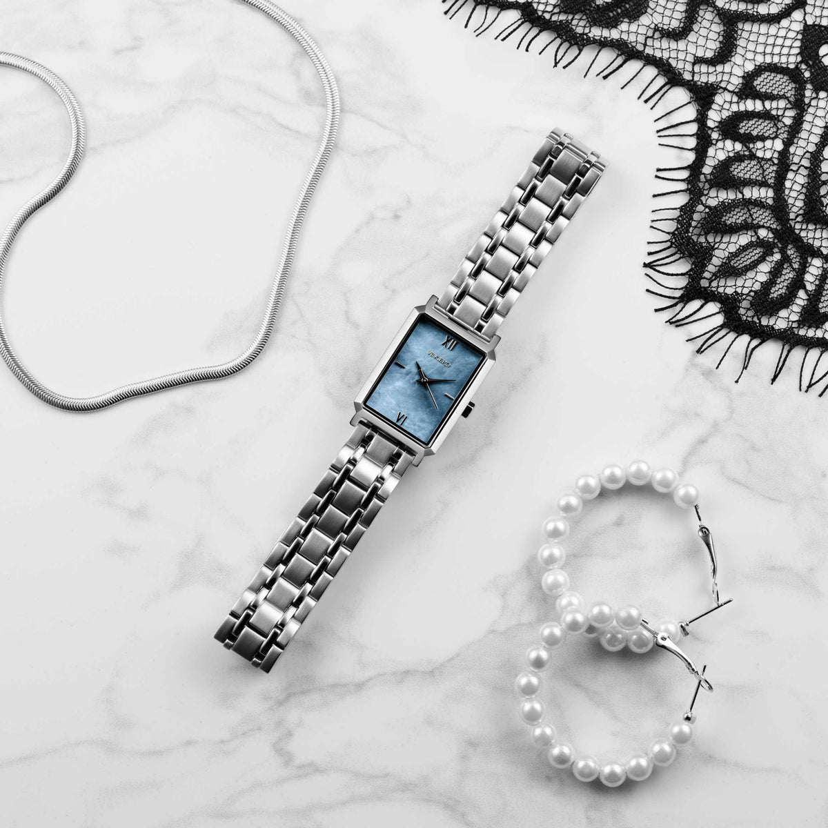 Silver watch with blue face on a marble surface with a pearl bracelet and lace fabric.