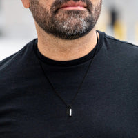 Man wearing a black necklace with a small pendant against a neutral background