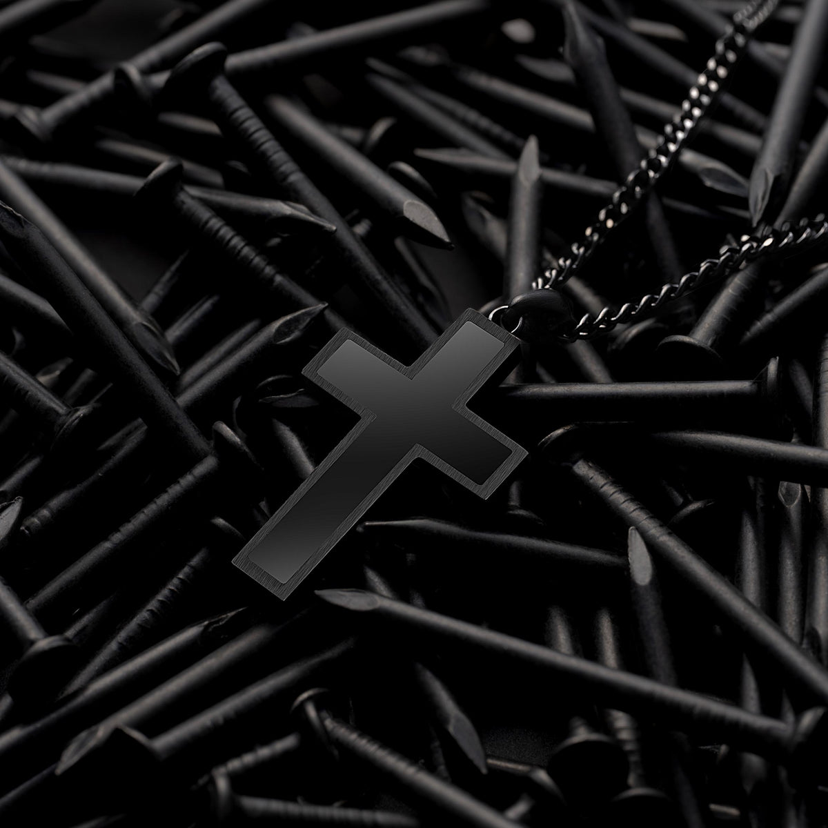 Close-up of a black cross pendant with a black chain, displayed on a pile of black nails.