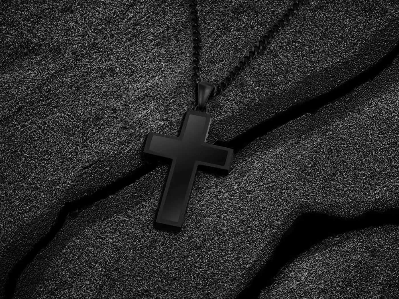 Close-up of a black cross pendant with a black chain, displayed on black sand. 
