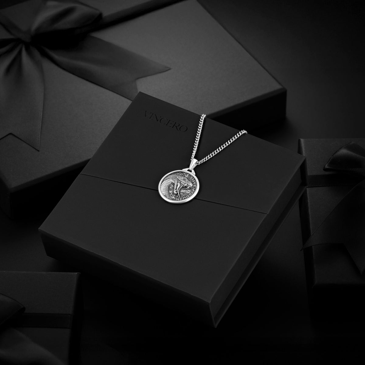 Silver necklace with a pendant on a black box, surrounded by black gift boxes.
