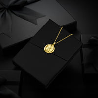 A gold circular pendant necklace with an engraved religious design, displayed on a black gift box with a black ribbon bow against a dark background.