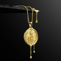 A gold pendant necklace with a circular coin-style charm featuring a rabbit motif and raised text, hanging from a gold chain against a black background with a dark textured ledge above.