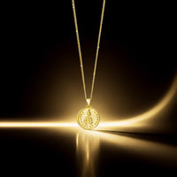 A gold pendant necklace with a circular coin-style charm featuring a rabbit motif and engraved text, suspended from a delicate gold chain against a dark background with dramatic warm light effects.