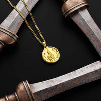 A gold chain necklace with a round, coin-style pendant featuring an embossed figure and text, displayed on a black background with rustic metal props.