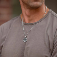 A close-up of a man wearing a silver chain necklace with a circular pendant, paired with a taupe-gray t-shirt.