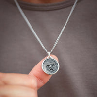 A close-up of a silver coin pendant necklace being held by a finger against a person's gray shirt. The pendant features an embossed design with a snake and the text 'SICILIA' and 'MILITUM'.