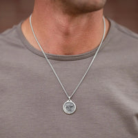 A silver chain necklace with a round, embossed pendant worn by a man in a gray t-shirt.