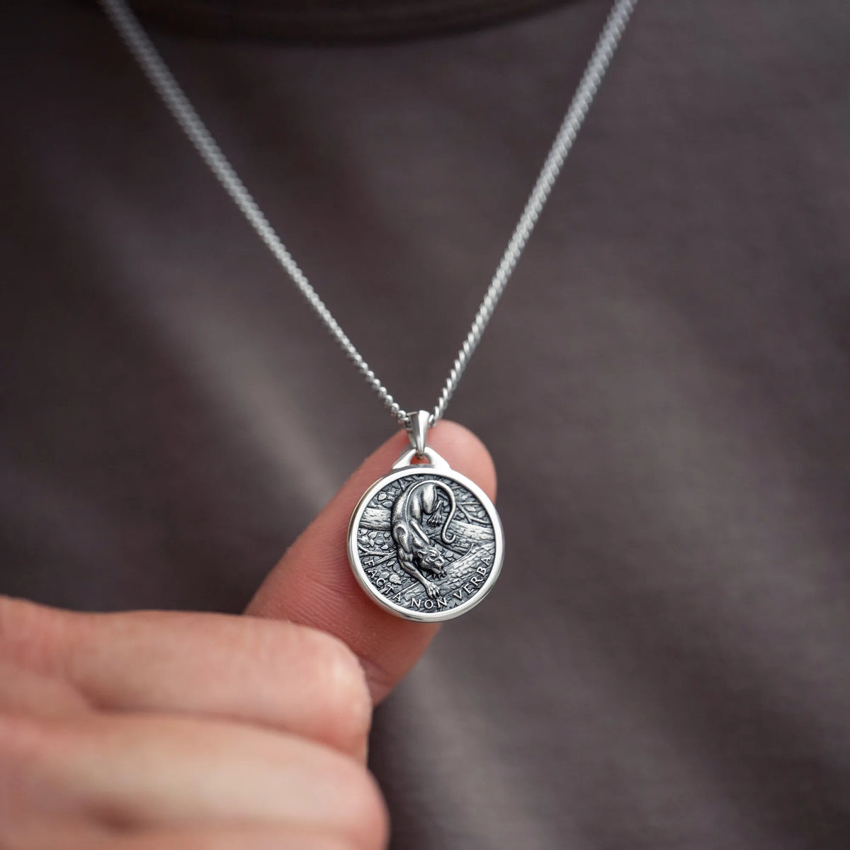 A close-up of a silver pendant necklace held by a finger against a dark gray shirt background. The pendant is circular with an embossed design of a panther figure.