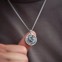 A close-up of a silver pendant necklace held by a finger against a dark gray shirt background. The pendant is circular with an embossed design of a panther figure.