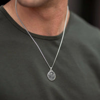 A close-up of a man wearing a silver chain necklace with an oval pendant featuring a skull and crossbones design, paired with a dark olive green t-shirt.