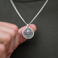 A close-up of a silver pendant necklace held by a hand against a dark gray shirt. The pendant is a circular coin-style charm featuring an eagle design and the text 'HONOR, SERVICE, DEDICATION'.