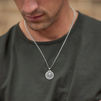 A silver chain necklace with a round, coin-style pendant featuring a classical bust, worn by a man in a dark olive green t-shirt.