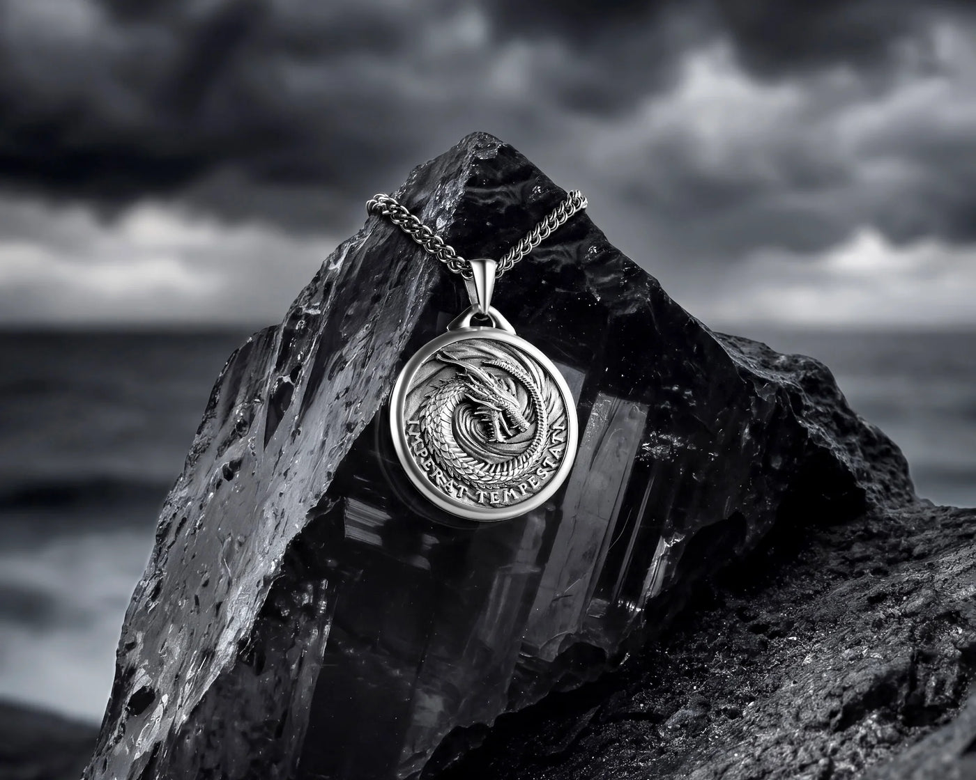 A silver-toned round pendant necklace with a dragon motif and chain, displayed on a dark rock with a blurred stormy seascape background.