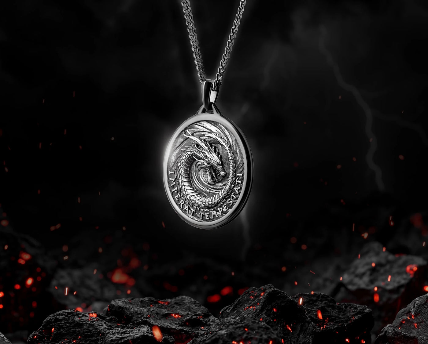 A silver oval pendant necklace featuring an embossed coiled dragon design, suspended from a matching chain against a dark, dramatic background with glowing embers and rocks.
