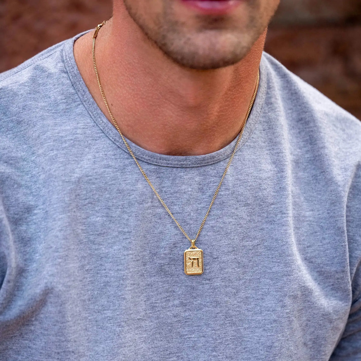 Man wearing a gold necklace with a rectangular pendant against a blurred background