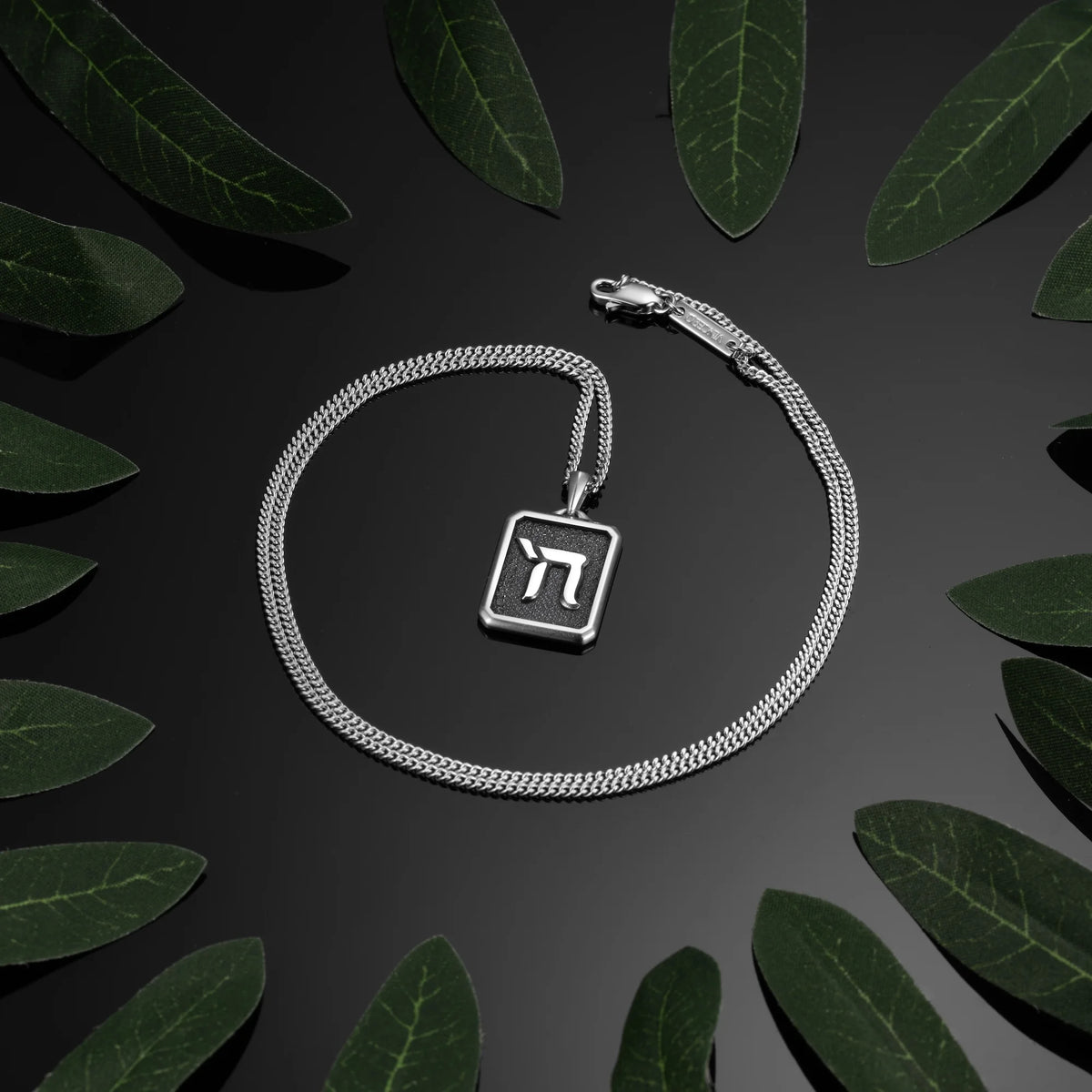 Silver necklace with a square pendant on a dark surface with green leaves