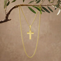A gold cross pendant necklace hanging from a gold chain, suspended from an olive branch against a warm beige background.