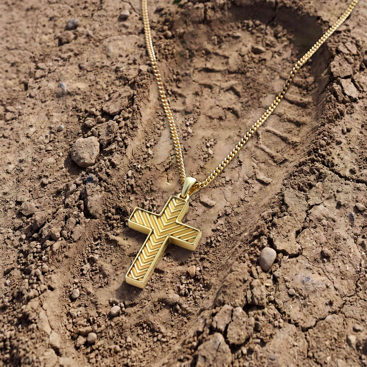 A gold cross pendant necklace with a textured chevron pattern, resting on a dry, cracked brown dirt surface.