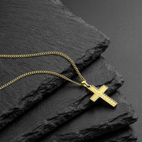 A gold cross pendant necklace with a textured chevron pattern on the cross and a matching chain, displayed on dark textured slate stones.