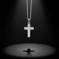 A silver cross pendant necklace with a textured chevron pattern, suspended from a silver chain against a dark background with a spotlight effect and a soft reflection below.