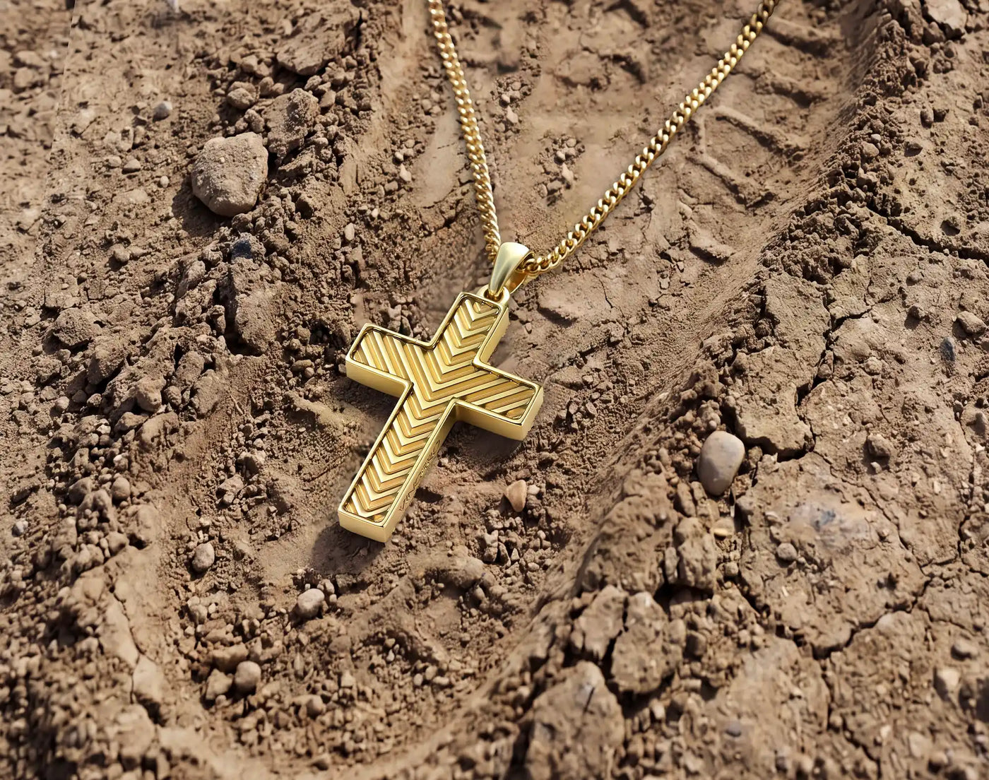 A gold cross pendant with a chevron pattern, hanging from a gold chain on a textured brown dirt background.