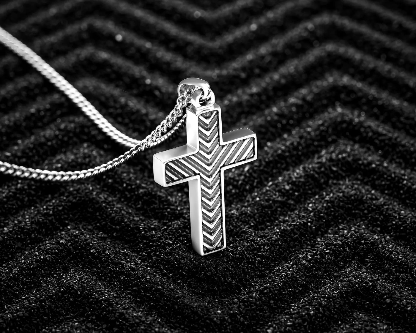A silver cross pendant necklace with a textured chevron pattern, resting on a black textured background.