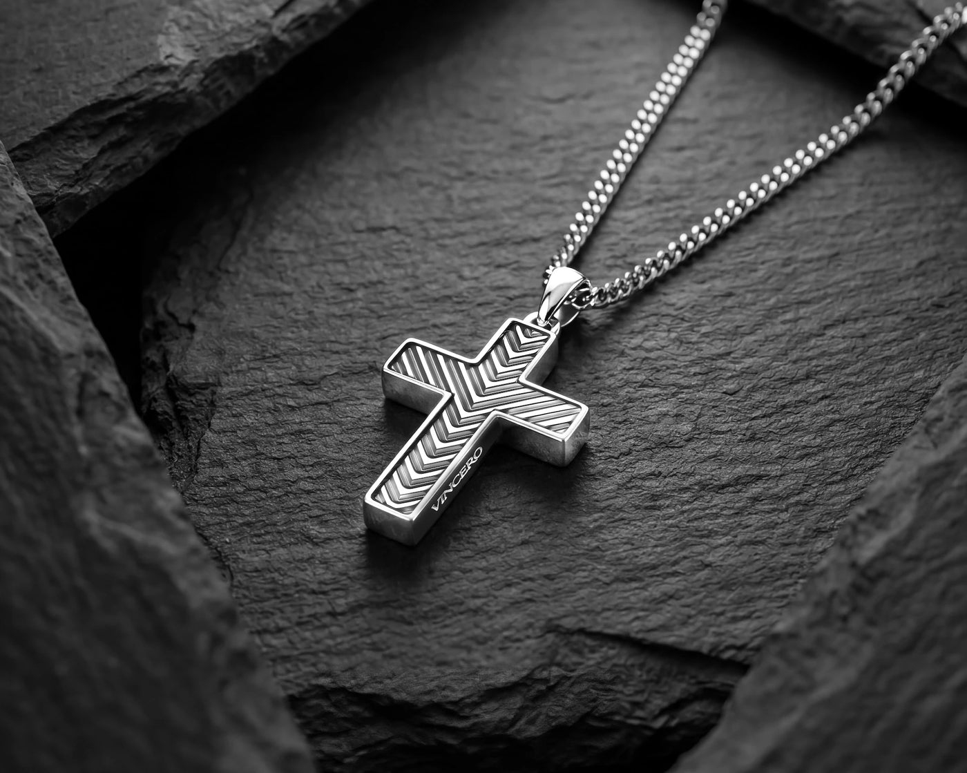 A silver cross pendant necklace with a textured chevron pattern, resting on a dark slate surface.