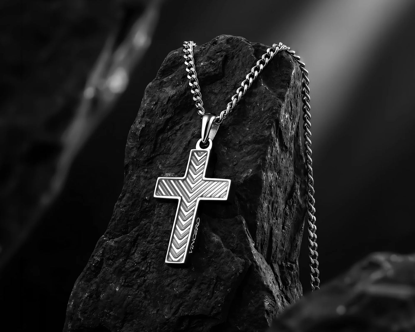 A silver cross pendant necklace with a chevron pattern, resting on a dark textured rock against a blurred black and white background.