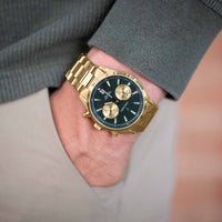 Gold wristwatch with a dark face on a person's wrist, wearing a gray suit.
