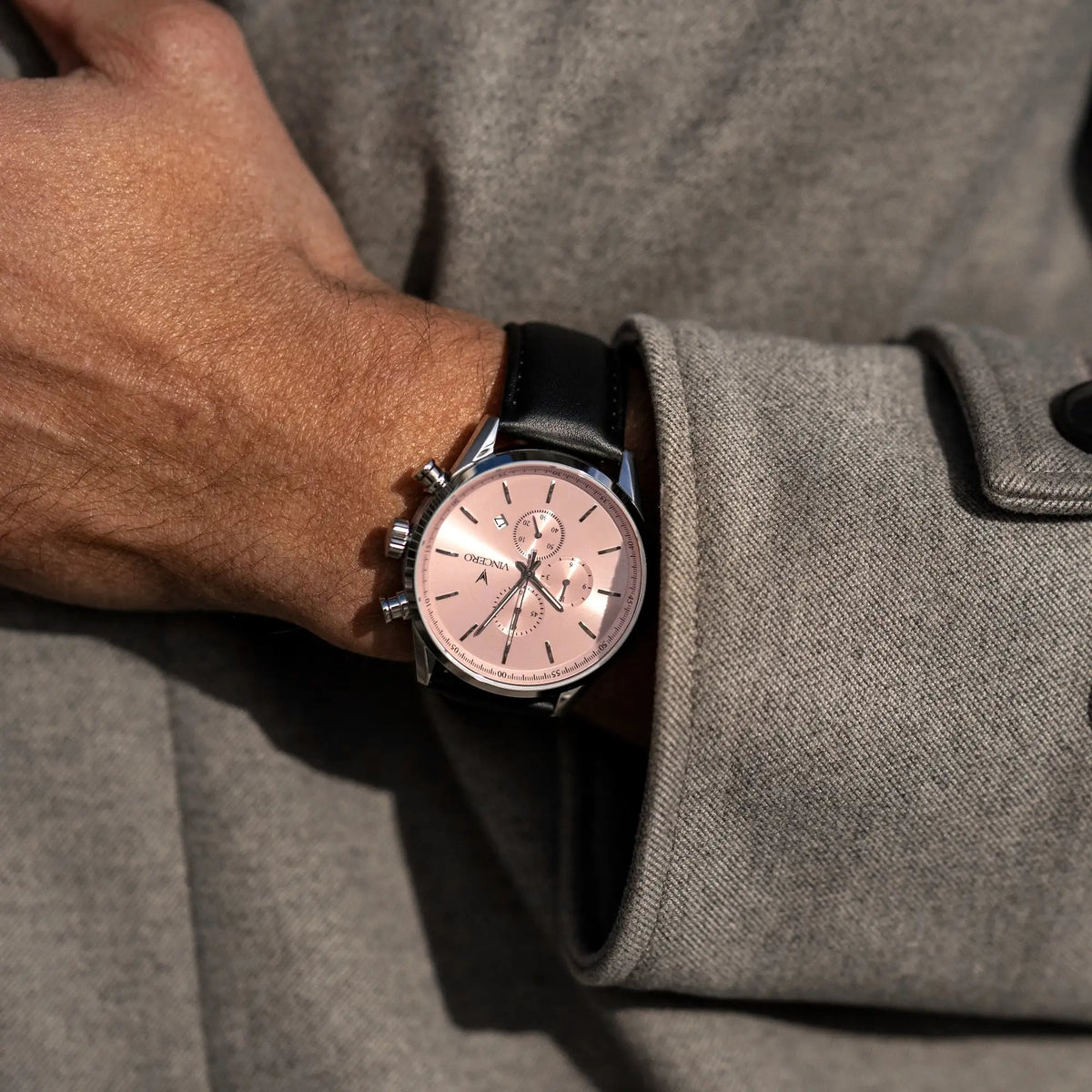 Close-up of a wrist wearing a watch with a pink face, set against a gray coat.