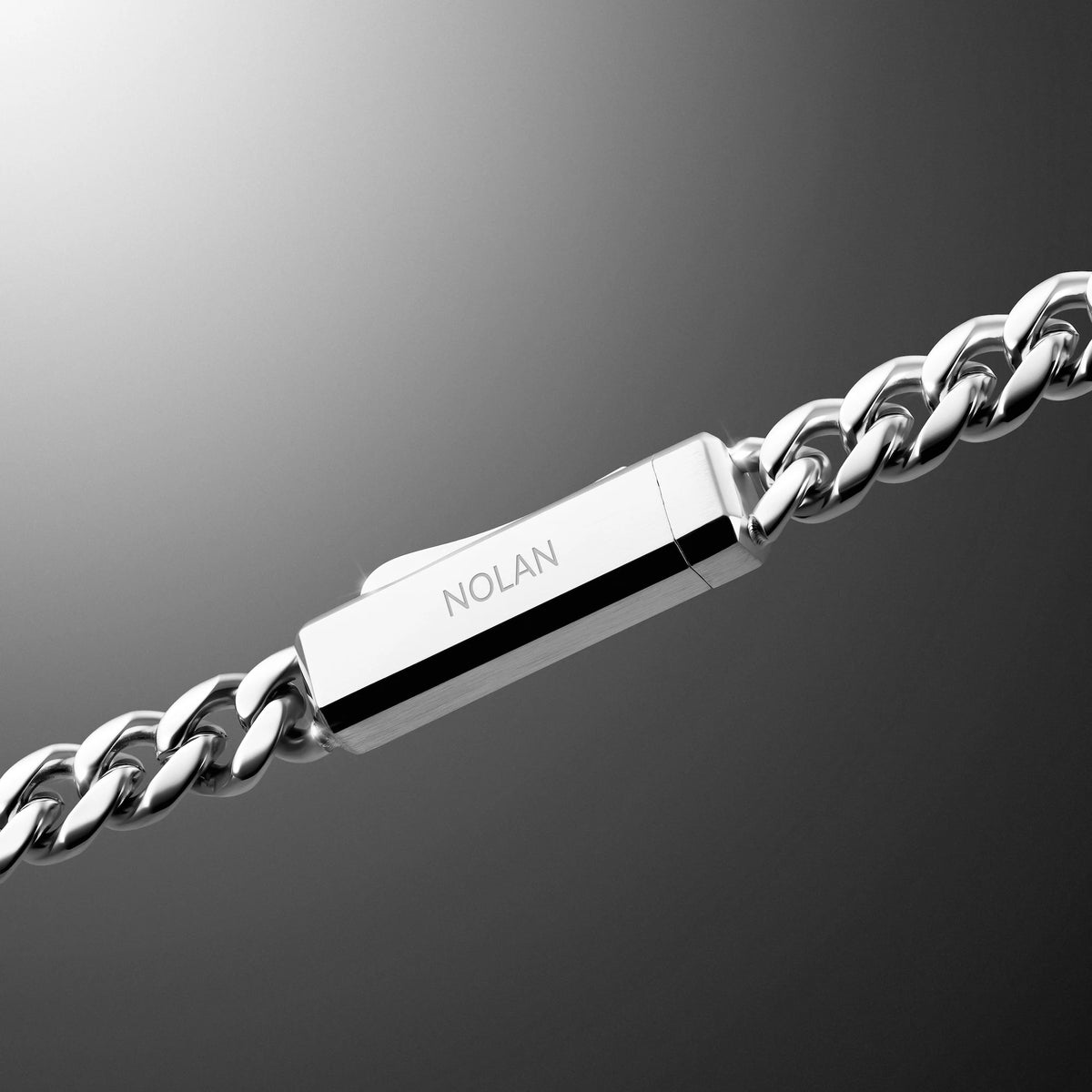 A close-up of a silver-toned metal chain bracelet with a rectangular clasp engraved with the brand name 'NOLAN', set against a dark gray gradient background.