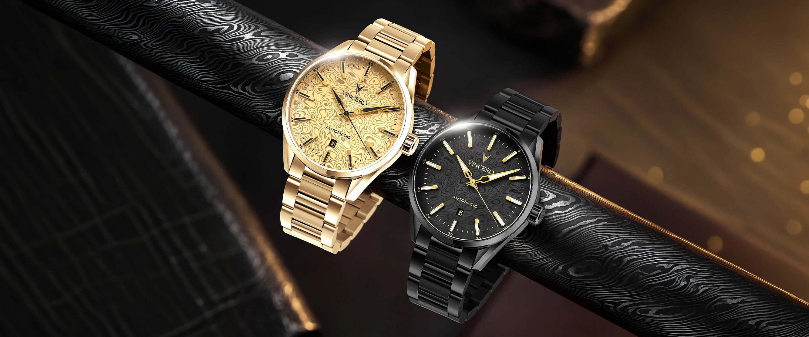 Two wristwatches displayed on a black textured strap against a dark warm-toned background. One watch has a gold-tone case and matching gold dial with a gold link bracelet.