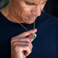 A close-up lifestyle shot of a man wearing a gold chain necklace with a green four-leaf clover pendant, holding the pendant between his fingers to show its details.