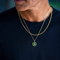 A close-up of a man's neck and lower face wearing a gold chain necklace with a green clover pendant over a dark navy blue t-shirt.