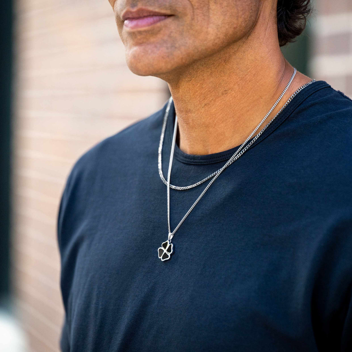 A close-up of a man wearing a layered silver necklace set with a geometric interlocking pendant, paired with a dark navy blue t-shirt against a blurred brick wall background.