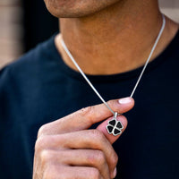 A close-up of a person wearing a silver chain necklace with a four-leaf clover pendant featuring black inlay, while holding the pendant with their hand.