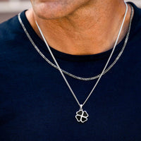 A close-up of a man's neck and chest wearing a silver chain necklace with a four-leaf clover pendant over a navy blue t-shirt.