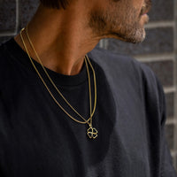 Man wearing a black shirt with a gold necklace featuring a clover pendant against a brick wall.