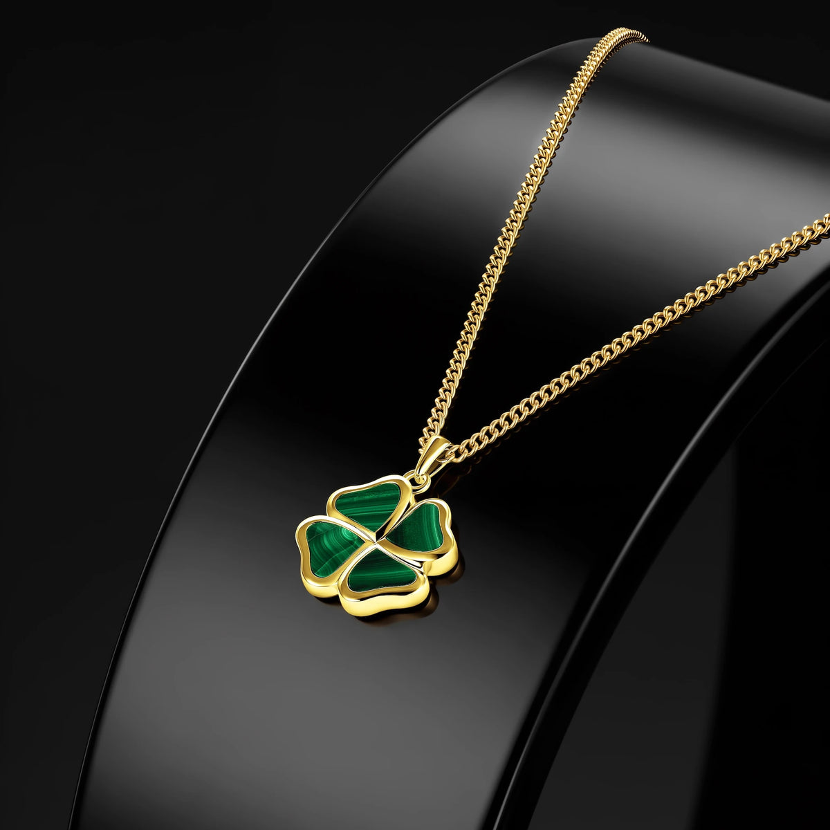 A gold chain necklace with a four-leaf clover pendant featuring green inlay, displayed on a glossy black jewelry bust against a dark background.