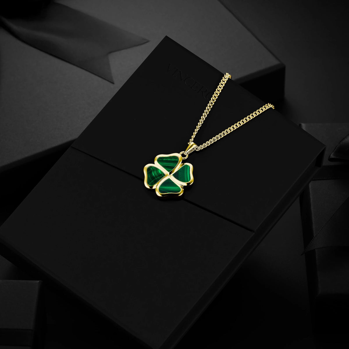 A gold chain necklace with a four-leaf clover pendant featuring green enamel inlay, displayed on a black gift box.