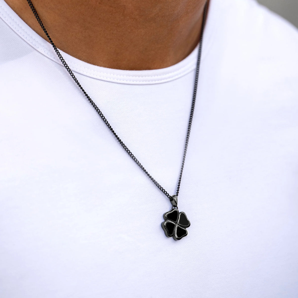 A close-up of a person wearing a black four-leaf clover pendant necklace on a black chain over a white t-shirt.