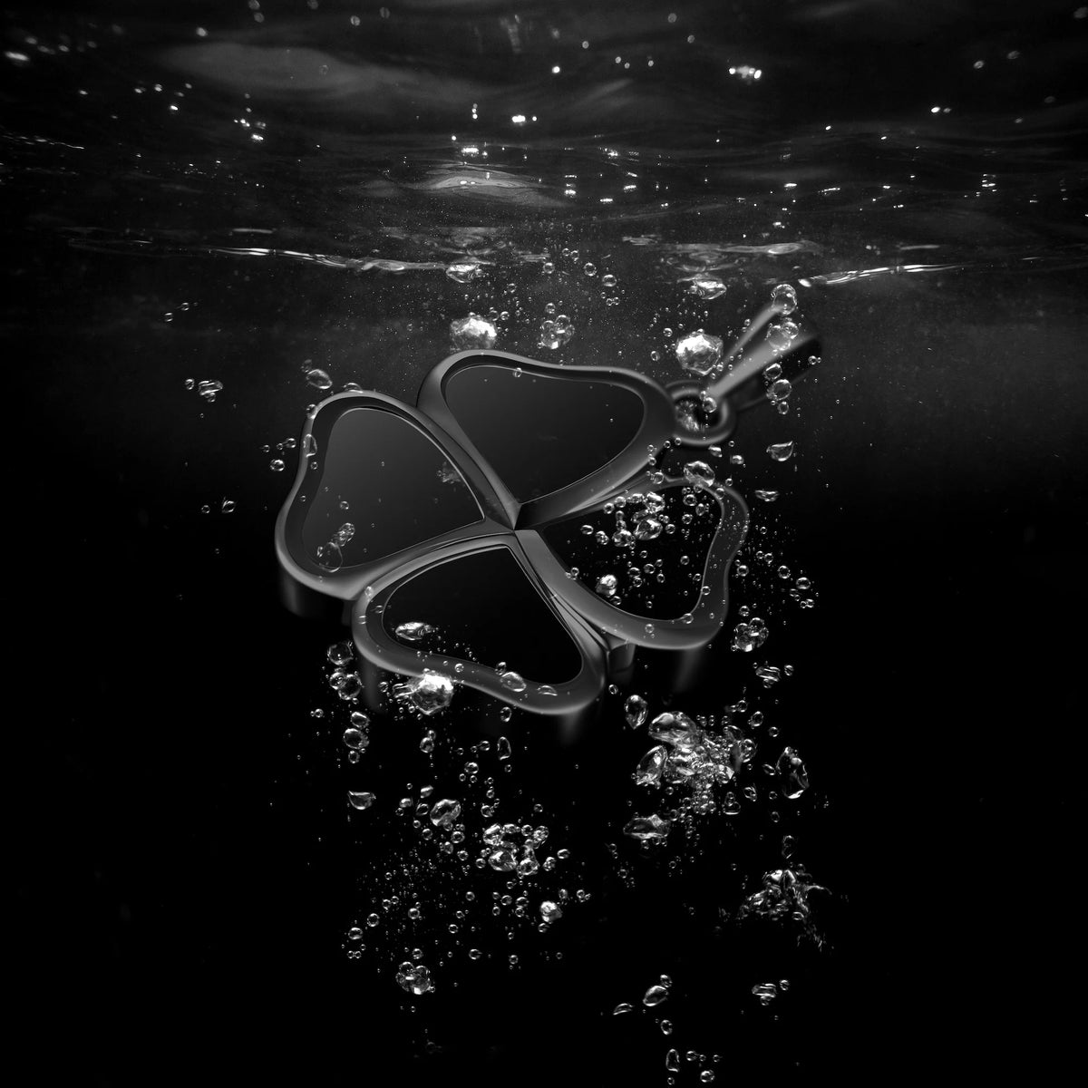 A dark metallic four-leaf clover pendant with a small hanging loop, floating underwater amidst rising bubbles against a black background.